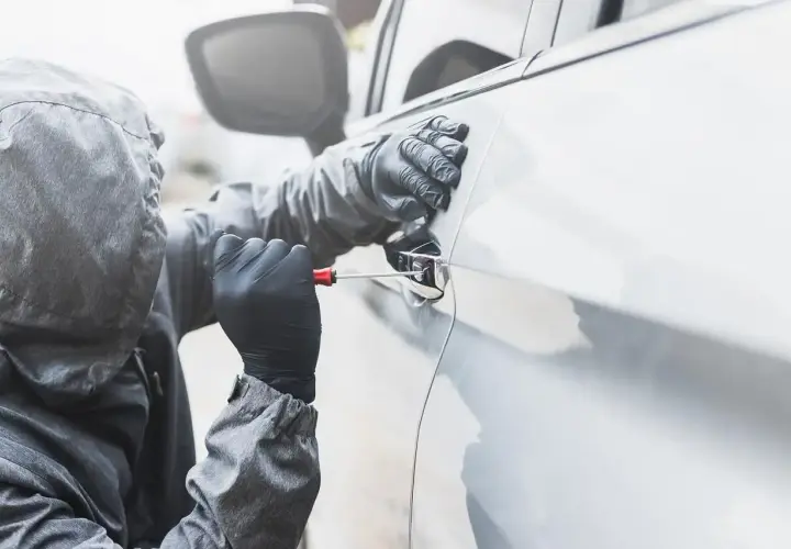 Osoba w kapturze i rękawiczkach próbuje otworzyć auto śrubokrętem. Sen o kradzieży auta może symbolizować utratę kontroli.