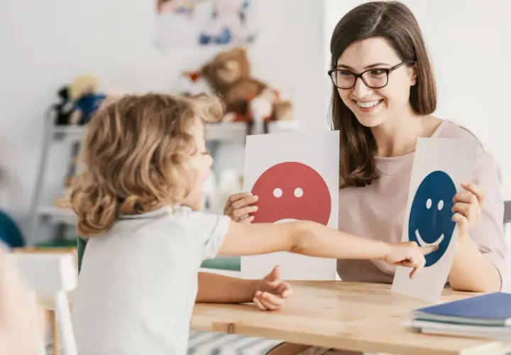 Dziecko wskazuje na uśmiechniętą buźkę, rozwijając inteligencję intrapersonalną. Terapeuta z uśmiechem prezentuje karty emocji.