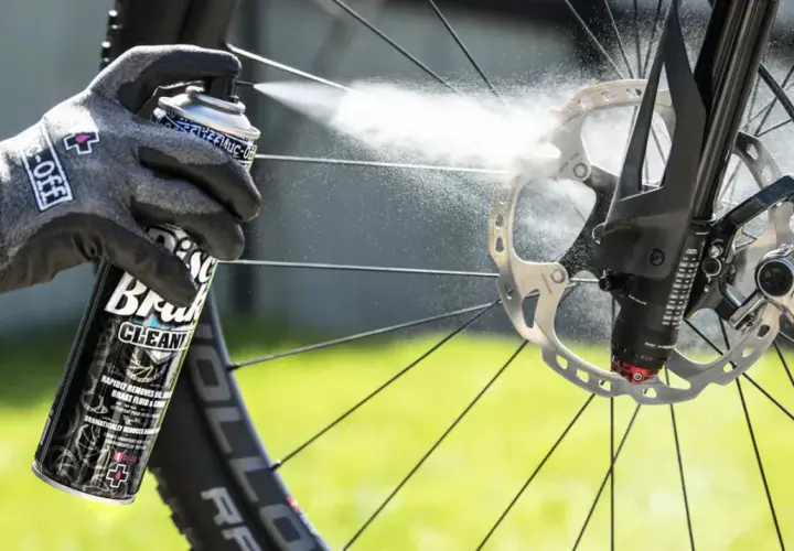 Gloved hand sprays Muc-Off Disc Brake Cleaner onto a bike's rotor, demonstrating how to clean up brake fluid for optimal performance.