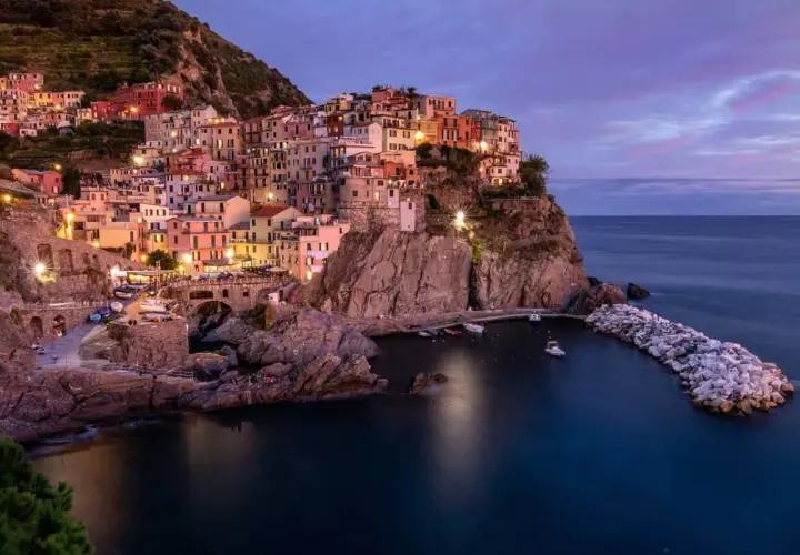 Manarola, una delle perle della strada panoramica 5 Terre, si illumina al crepuscolo. Case colorate aggrappate alla roccia, un piccolo porto e il mare blu intenso.
