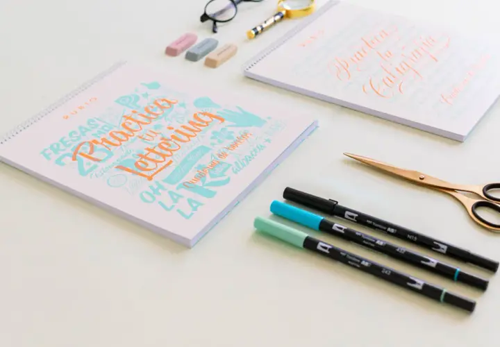 Cuadernos de "Practica Lettering" y "Practica tu Caligrafía" con rotuladores y tijeras doradas.