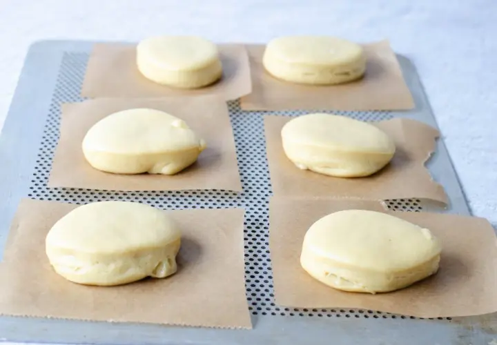 Seis bolas de fraile crudas, listas para hornear, descansan sobre papel de horno en una bandeja metálica.