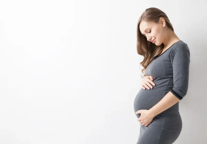 Uśmiechnięta ciężarna kobieta w szarym ubraniu delikatnie dotyka brzucha. To naturalne zmiany, jak rozstępy w ciąży, które są częścią tej wyjątkowej podróży.