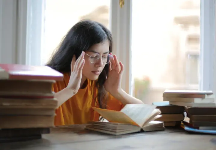 Joven con gafas y libros, con las manos en las sienes, buscando alivio al estrés con magnesio.