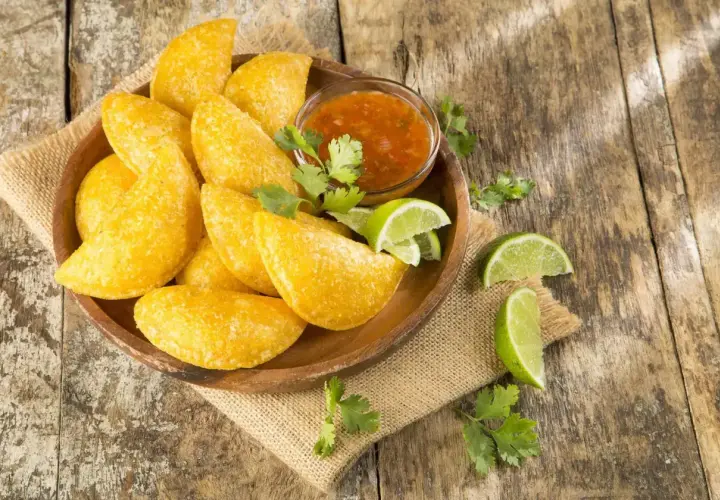 Empanadas colombianas doradas en plato de madera con salsa, limones y cilantro sobre mesa rústica.
