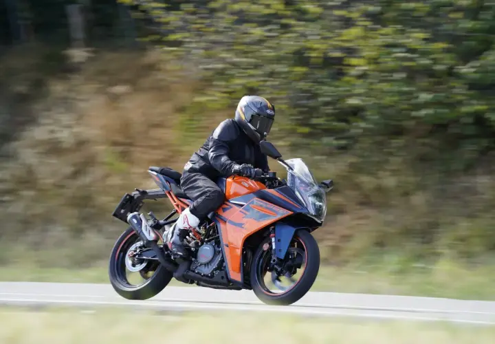 Motociclista con casco y traje negro conduce una KTM RC 390 CV naranja y azul por una carretera con árboles borrosos al fondo.
