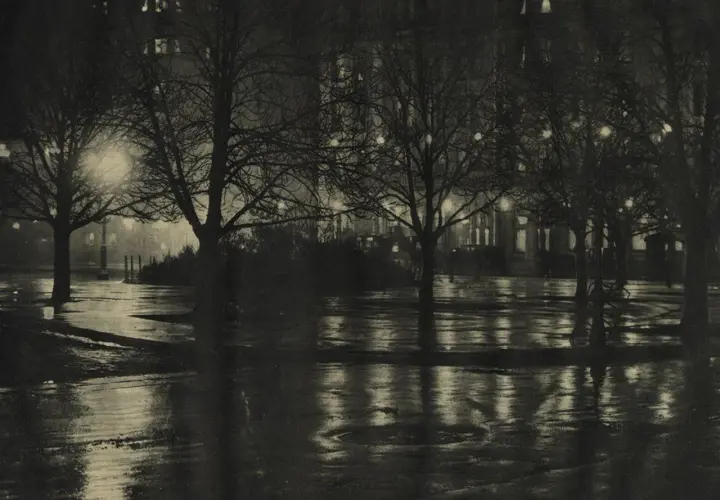 Nocny park, mokre alejki odbijające światła latarni i okien. Piktorializm w fotografii oddaje nastrój i atmosferę.