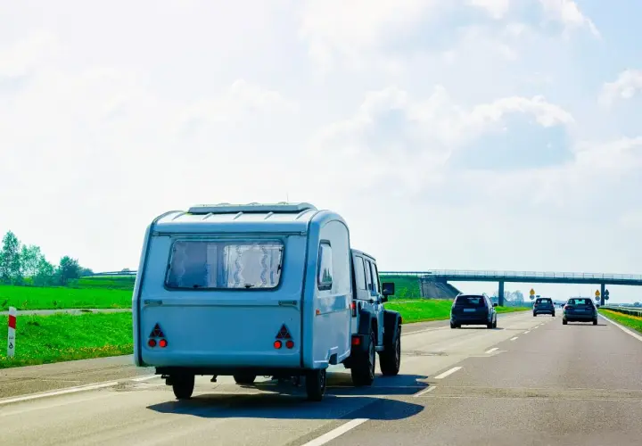 Samochód terenowy ciągnie dmc przyczepy kempingowej po autostradzie. W tle zielone pola i most.