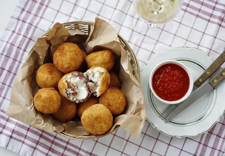 Koszyk pełen złotych arancini, jedno przekrojone ukazuje ryż i ser. Obok miseczka z sosem pomidorowym. Idealne na przekąskę, gdy masz ochotę na arancini przepis.