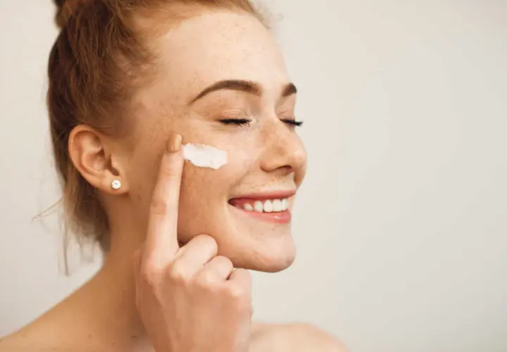 Une jeune femme rousse souriante, les yeux fermés, applique de la crème sur sa joue. Elle montre comment appliquer crème visage pour une peau éclatante.