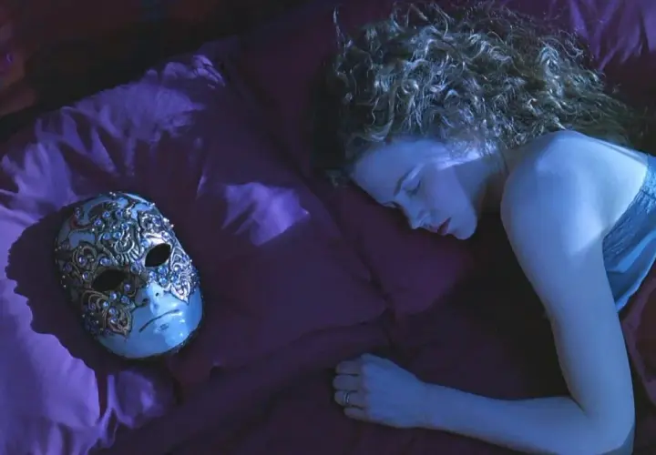 Una donna dorme su un letto di seta viola, accanto a una maschera veneziana ornata. La sua curiosità è palpabile, come se stesse sognando occhi chiusi.