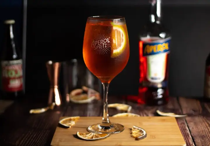 Orzeźwiający drink z aperolem, udekorowany plasterkiem pomarańczy, stoi na drewnianej desce. W tle butelka aperolu i inne akcesoria barmańskie.