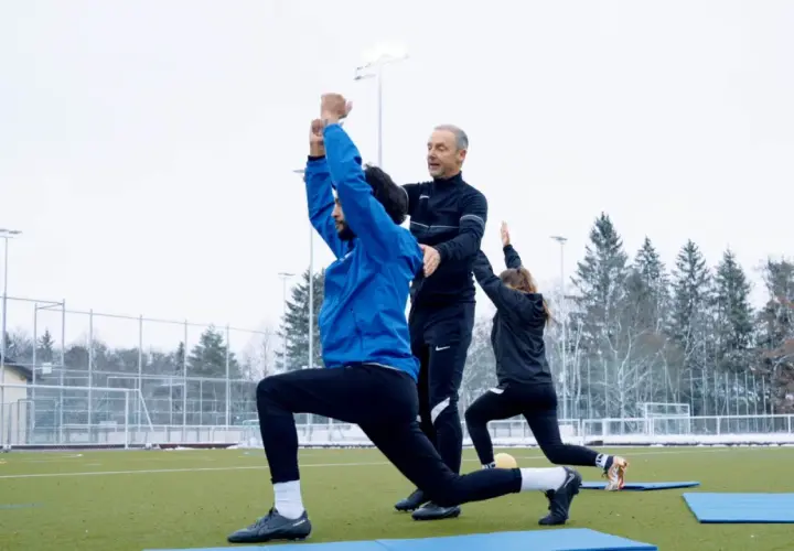 Athletik Training Fußball: Trainer korrigiert Ausfallschritt bei Spieler auf Kunstrasen. Eine Spielerin macht mit.