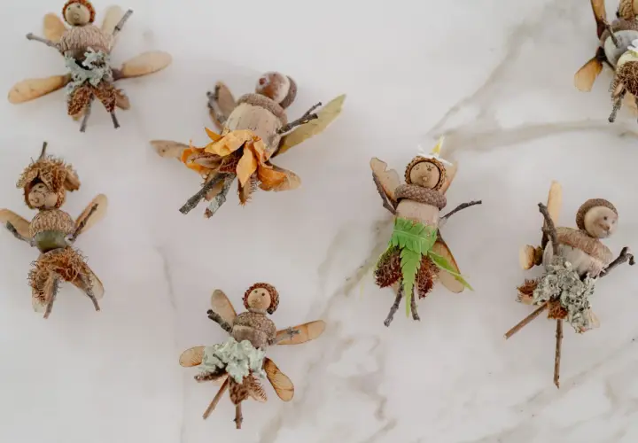 Figuras de hadas hechas con bellotas, ramas y hojas sobre fondo blanco.