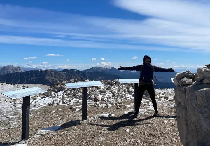 Persona amb els braços oberts al cim del Pedra dels Quatre Batlles, amb paisatge muntanyós i cel blau.