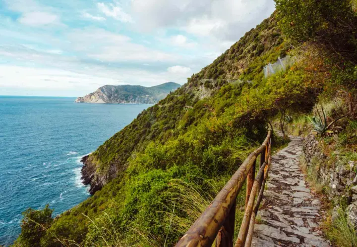 A stone path winds along the coast for Cinque Terre hiking, with lush green hills and the blue sea stretching to the horizon.