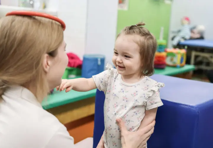 Uśmiechnięte dziecko w kwiecistym body wyciąga rękę do terapeuty w gabinecie.