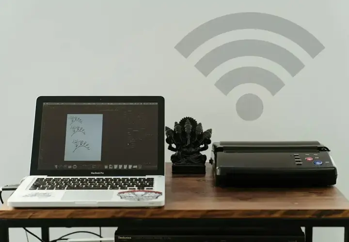 Laptop, drukarka i figurka Ganesha na biurku. Ikona Wi-Fi na ścianie sugeruje, jak połączyć drukarkę z wifi bezprzewodowo.