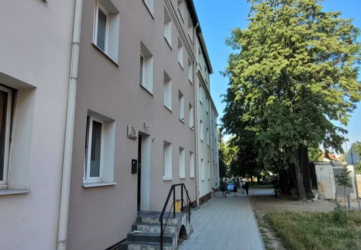 Budynek mieszkalny z oknami i drzwiami, obok drzewa i chodnik. To miejsce może być związane z art. 18 ustawy o ochronie praw lokatorów.