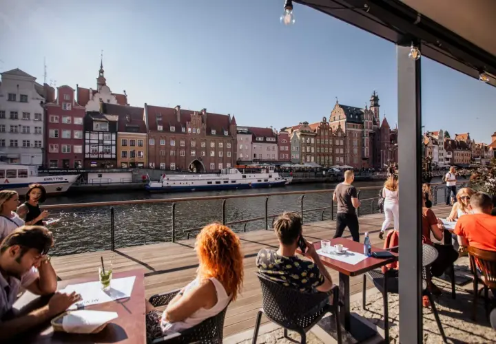 Gdańsk restauracje z widokiem na Motławę. Ludzie siedzą przy stolikach na drewnianym tarasie, podziwiając zabytkową zabudowę i pływające statki.