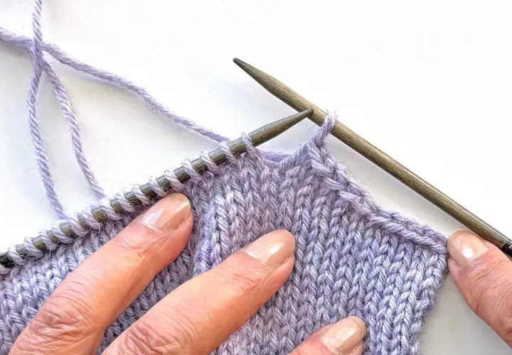 Close-up of hands sewing shoulder seams in knitting with lilac yarn and wooden needles.
