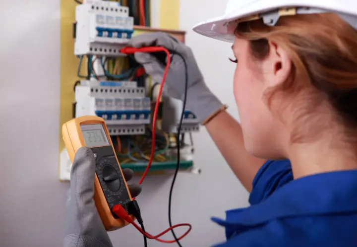 Elektryk w białym kasku wykonuje pomiary elektryczne przy skrzynce z bezpiecznikami, trzymając multimetr.