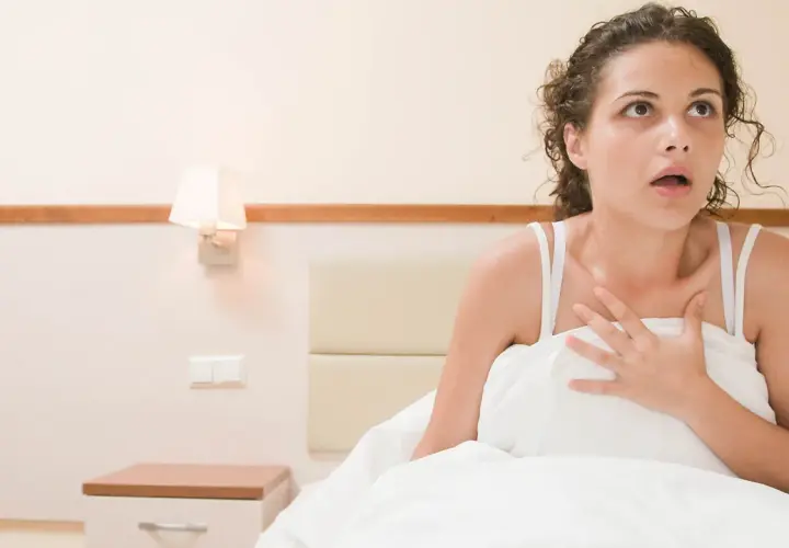 Mujer despierta en cama, con expresión de asombro y mano en el pecho, como si acabara de tener un sueño vívido: soñar que mi padre fallecido está vivo y me habla.