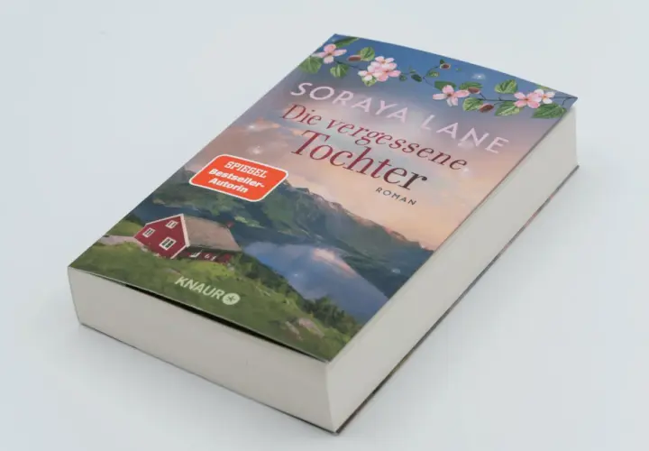 Ein Buchcover mit dem Titel "Die vergessene Tochter" von Soraya Lane. Es zeigt eine rote Hütte am See vor einer Bergkulisse.
