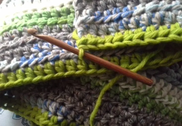 Coperta ai ferri con avanzi di lana, un'opera in corso con un uncinetto di legno che lavora maglie verdi su uno sfondo multicolore.
