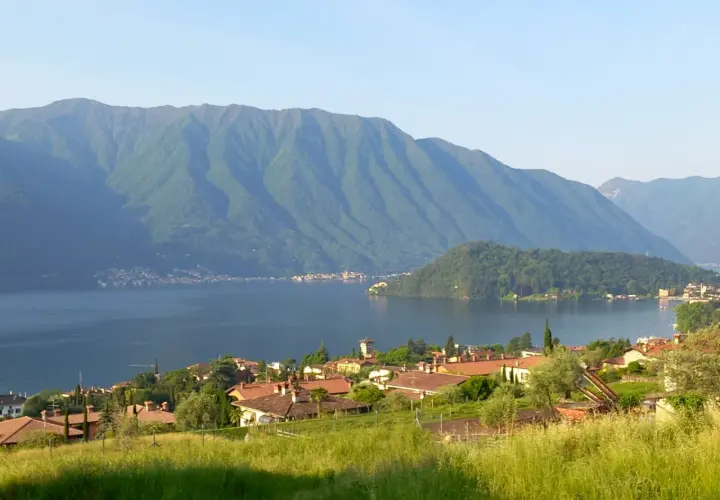 Villaggio sulle rive del lago più grande d'Europa, con montagne verdi sullo sfondo e un prato fiorito in primo piano.