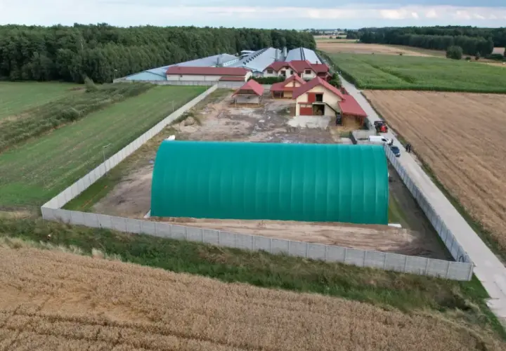Zielona hala namiotowa na terenie zabudowy siedliskowej, otoczona polami i lasem.