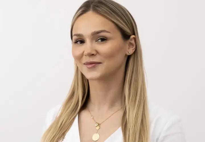 Kobieta z blond włosami i delikatnym makijażem, uśmiecha się lekko, choć jej nienaturalny wyraz twarzy sugeruje, że pozuje do zdjęcia.