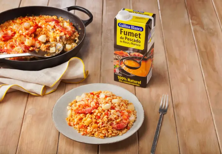 Arroz meloso con gambas y trozos de pescado en paellera y plato, junto a un cartón de fumet.