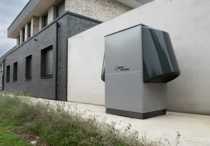 Nowoczesna pompa ciepła, szara, z logo "alpha innotec", stoi przy betonowej ścianie obok budynku z cegły.
