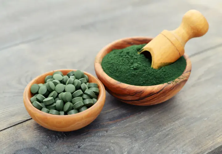 Due ciotole in legno: una con compresse verdi e l'altra con polvere di **pianta alga spirulina**, con un cucchiaino di legno.