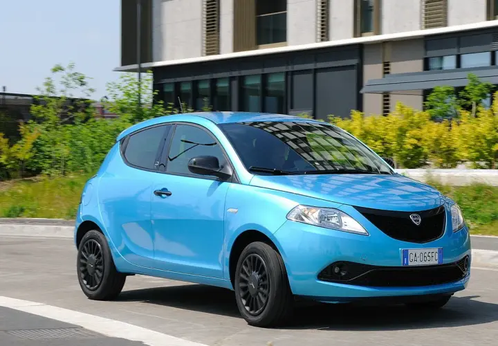 Lancia Ypsilon 1.3 Multijet Diesel azzurra in movimento, con sfondo di edifici moderni e vegetazione.