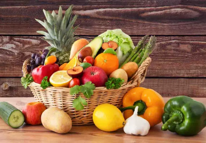 Cesto pieno di frutta e verdura fresca: ananas, uva, mele, arance, limoni, pomodori, peperoni, patate, cetrioli, asparagi e lattuga.