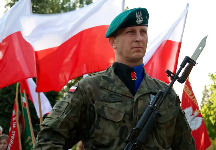 Żołnierz w zielonym beretcie, z karabinem i bagnetem, na tle dwóch biało-czerwonych flag.