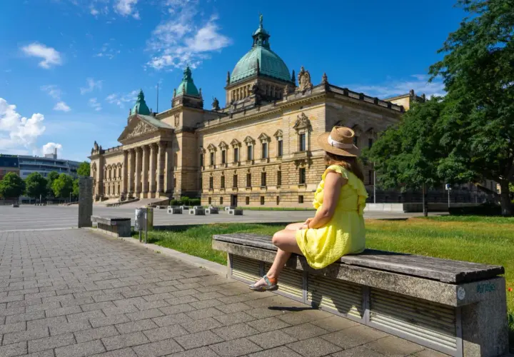 Kobieta w słomkowym kapeluszu i żółtej sukience siedzi na ławce przed monumentalnym budynkiem w Lipsku, mieście w Saksonii znanym z międzynarodowych targów.