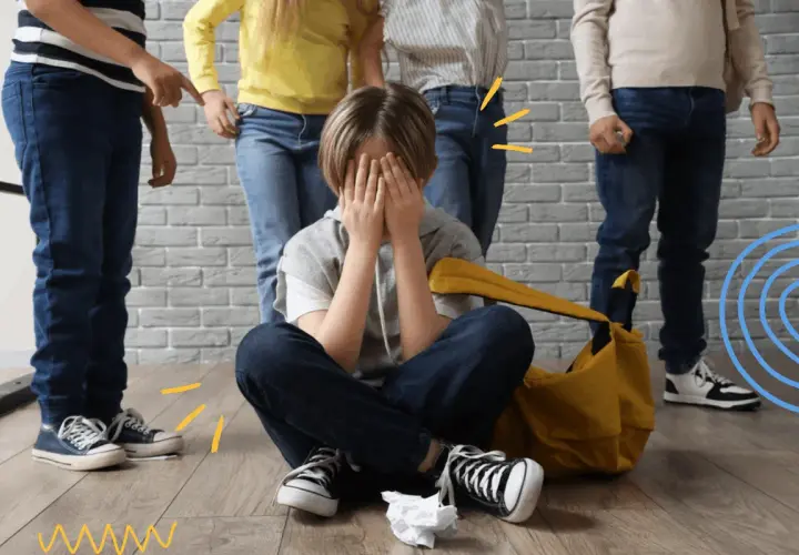 Chłopiec siedzi na podłodze, zakrywając twarz dłońmi, podczas gdy inne dzieci wskazują na niego. To obraz tego, czym jest bullying.