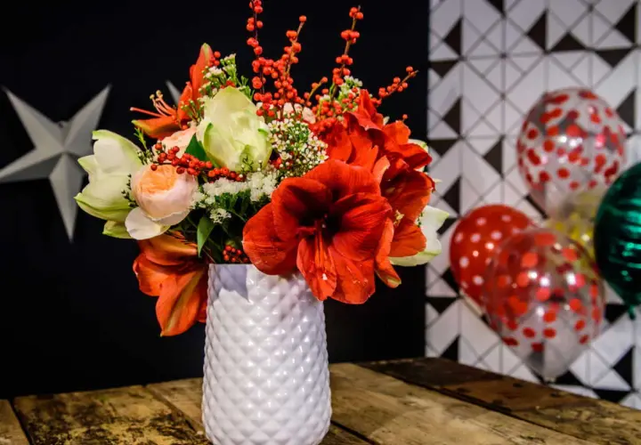 Magnifique composition amaryllis rouge et blanche, ornée de baies rouges, dans un vase texturé. Ballons festifs en arrière-plan.
