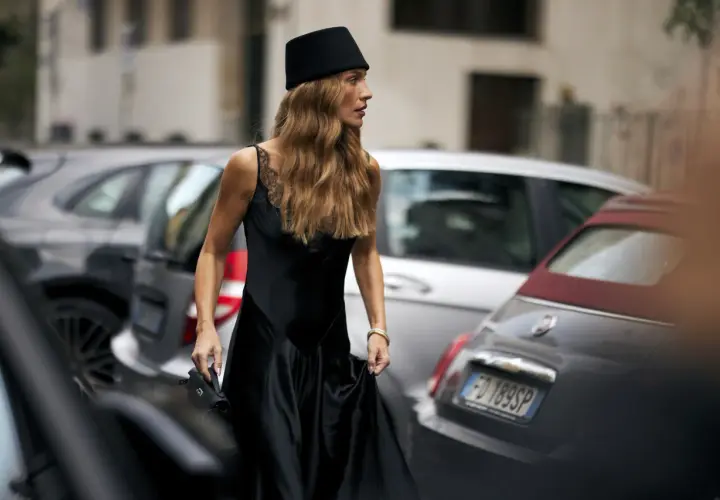 Mujer con sombrero negro y vestido de seda, su pelo en pausa, camina entre coches aparcados.