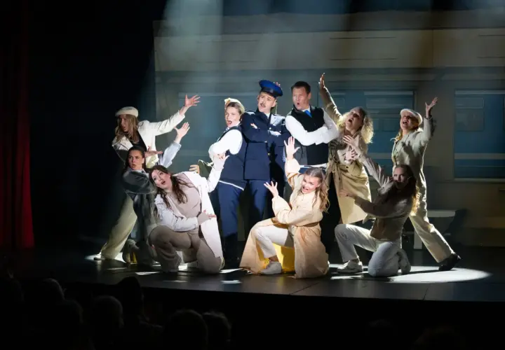 Ensemblen i en svensk revy, med en polisman i centrum, gestikulerar dramatiskt på scenen framför en tågvagnsbakgrund.