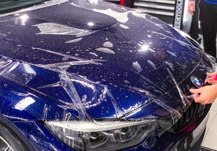 Nakładanie folii ochronnej na maskę ciemnoniebieskiego BMW. Widać krople wody i pęcherzyki powietrza pod folią.
