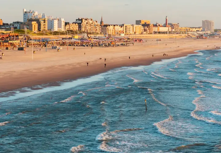Ein sonniger Tag an der Nordsee Holland. Wellen brechen am Strand, im Hintergrund die Stadtkulisse mit Hotels und Strandpavillons.