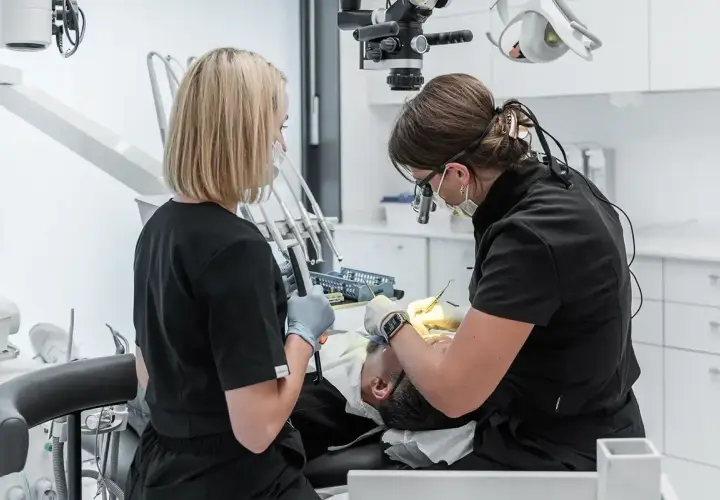Dwie dentystki pracują przy pacjencie. Jedna używa mikroskopu, druga trzyma narzędzie. To obrazek o tym, jak zostać dentystą.