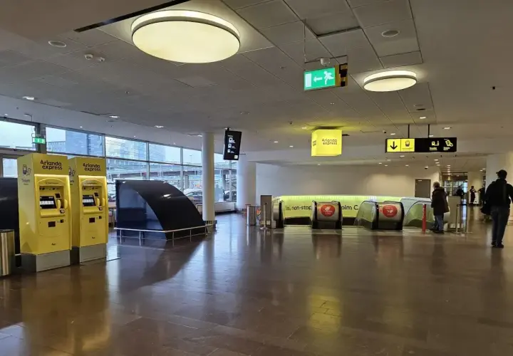 Żółte automaty z biletami na Arlanda Express na sztokholmskim lotnisku. W tle schody ruchome i znaki kierunkowe.