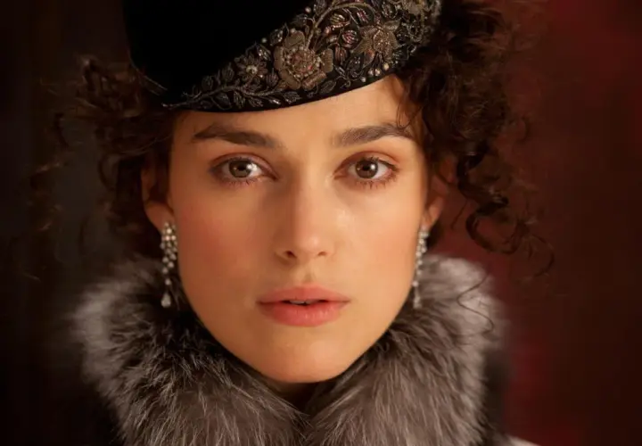 Anna Karenina w eleganckim futrze i ozdobnym kapeluszu, z błyszczącymi kolczykami, patrzy wprost przed siebie.