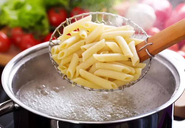 Gotowy do gotowania makaron penne w cedzaku nad bulgoczącym garnkiem. W tle pomidory i sałata.