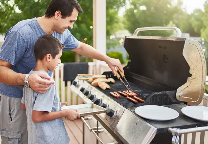 Un père et son fils préparent des saucisses sur un barbecue à gaz. Le père utilise des pinces pour retourner les saucisses pendant que le fils tient un plateau.